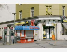 Ein Stadtbild mit einer gelb-grünen Gebäude-Ecke, darauf das Logo und der Eingang einer Commerzbank-Filiale. Davor steht ein kleiner geschlossener Imbissstand mit der Aufschrift "Ayaz Grill" und zwei öffentliche Telefonzellen. Ein gelbes Haltestellenschild und ein oranger Briefkasten sind ebenfalls sichtbar.