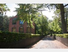 Backsteinhaus mit grünen Fensterrahmen und dekorativem Giebeldetail, umgeben von Bäumen und einer schmalen Kiesstraße, die von Hecken und Holzzäunen gesäumt ist. Der Ort liegt in einer waldreichen, ruhigen Umgebung.