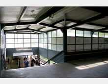 Überdachter Zugang zu einem Bahngleis mit Betonboden und Metallgeländern, umgeben von Milchglasfenstern. Im Hintergrund führt eine Treppe hinunter zu den Gleisen, wo ein Schild mit den Richtungen „Oranienburg“ und „Berlin-Wannsee“ zu sehen ist.