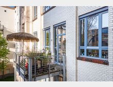 Mehrstöckiges Gebäude mit hellen Klinkerfassaden, großen blauen Fenstern und einem kleinen Balkon aus Holz und Metallgeländern. Der Balkon ist mit einer Strohdach-Sonnenschirm, Pflanzen, Stühlen und einem kleinen Tisch ausgestattet.