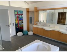 Modernes Badezimmer mit grauem Fliesenboden, Doppelwaschbecken mit Holz-Unterschränken und großem Wandspiegel. Im Vordergrund steht eine weiße Badewanne, während an der Wand ein abstraktes Gemälde und dekorative Poufs sichtbar sind.