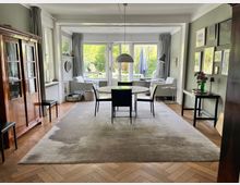 Wohnzimmer mit einer offenen, hellen Gestaltung. Im Vordergrund steht ein runder Esstisch mit Stühlen auf einem großen Teppich, dahinter eine Sitzecke mit zwei Sesseln vor bodentiefen Fenstern, die einen Blick auf einen Garten bieten.