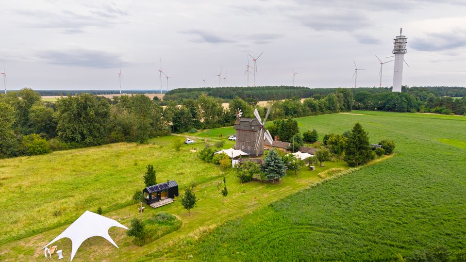 Luftaufnahme einer ländlichen Landschaft mit Feldern und Baumgruppen, in deren Mitte eine historische Windmühle mit Nebengebäuden und weißen Zeltdächern steht. Am Horizont sind mehrere Windkraftanlagen sowie ein hoher Aussichtsturm am Waldrand zu sehen.