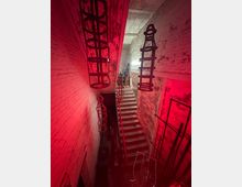 Ein schmaler, industriell wirkender Treppenaufgang in einem Gebäude mit weißen, teils verwitterten Ziegelwänden, beleuchtet von roten Lichtern. Metallstrukturen hängen von der Decke, und der Raum wirkt wie ein Lager- oder Werkstattbereich.