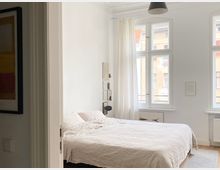Helles Schlafzimmer mit weißem Doppelbett und hohen, weiß gerahmten Fenstern mit transparenten Vorhängen. Weiße Wände mit wenigen Bildern, eine schwarze Deckenlampe und ein heller Holzboden mit Teppich prägen den Raum.