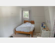 Schlichtes Schlafzimmer mit weiß gestrichenen Wänden, einem Einzelbett mit Holzrahmen und einfacher Bettwäsche. Neben dem Fenster mit floralen Vorhängen steht ein schmaler weißer Schrank, auf dem verschiedene persönliche Gegenstände liegen.