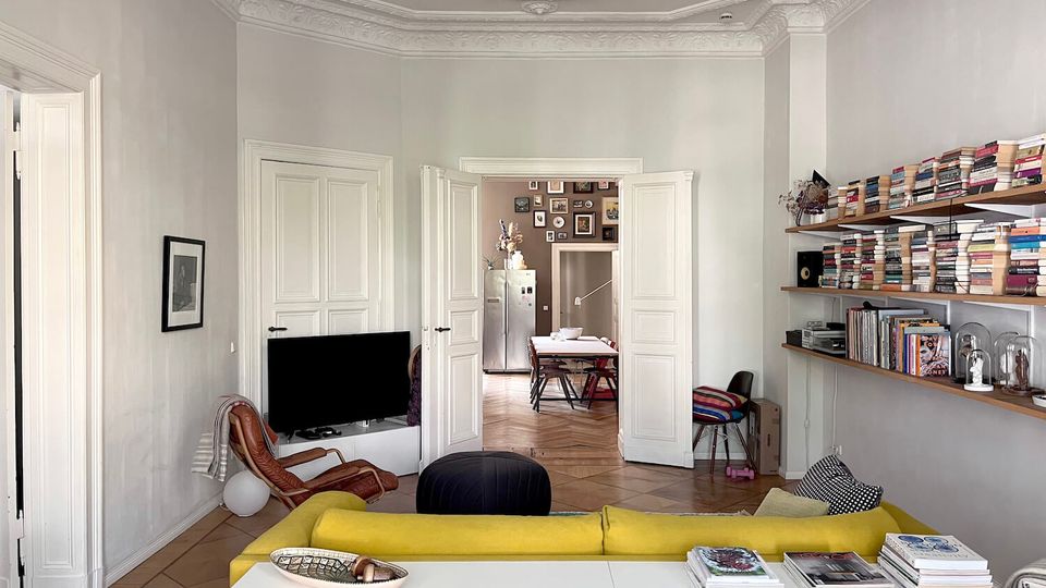 Bright, high-ceiling living room with ornate plaster crown molding, white walls, and a tiled floor, furnished with a yellow sofa, low white media console, and a TV. Long wall-mounted shelves filled with books line the right wall, and open double doors lead to a dining area with a table and wall gallery beyond.