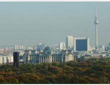 Der Blick zeigt das Berliner Stadtzentrum mit dem Reichstagsgebäude im Vordergrund, erkennbar an seiner gläsernen Kuppel, und dem Fernsehturm im Hintergrund. Im unteren Bereich dominieren die herbstlich gefärbten Bäume des Tiergartens die Szene.
