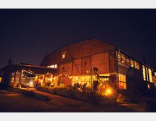 Ein großes, altes Backsteingebäude mit zahlreichen Fenstern, das bei Nacht von warmem Licht beleuchtet ist. Im Vordergrund befindet sich ein überdachter Außenbereich mit Lichterketten und Dekoration, umgeben von Pflanzen und kleinen Sitzgruppen.