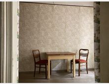 Ein einfacher Innenraum mit einem Holztisch und zwei Holzküchenstühlen mit roten Polstern in der Mitte des Raumes, umgeben von beigefarbener Tapete mit floralem Muster; links und rechts im Vordergrund sind geöffnete Türen mit gemusterten Tapeten sichtbar.