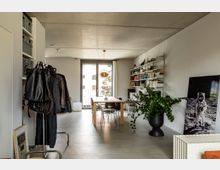 Minimalistisch eingerichteter Wohnraum mit Betonboden und heller Wandfarbe. Im Vordergrund ein Kleiderständer und eine große Zimmerpflanze, im Hintergrund ein Esstisch aus Holz, daneben ein Regal mit Büchern, ein großes Fenster mit Blick nach draußen und eine goldene Hängelampe.