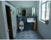 Modernes Badezimmer mit weißen Wänden und grauen Fliesenböden. Sichtbar sind ein Wandwaschbecken mit Spiegel, ein Toilettebereich mit einer Glasabtrennung, ein großes Fenster mit einem Holzeimer auf der Fensterbank und an der Wand hängende gelbe Handtücher.