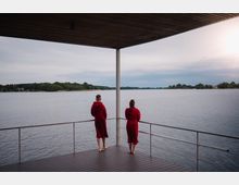 Zwei Personen in roten Bademänteln stehen auf einer überdachten Holzterrasse mit Geländer und blicken auf einen ruhigen See, der von bewaldeten Ufern und einem wolkigen Himmel umgeben ist.