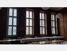 Ein Innenraum mit dunklen Holzwänden und einer Reihe großer Fenster, die mit dekorativen Gittern versehen sind. Im Vordergrund stehen einfache Holztische und Plastikstühle, während von außen etwas Tageslicht hereinfällt.