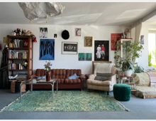 Cozy living room with a brown tufted sofa, a striped armchair, a glass coffee table on a large green rug, and a tall wooden bookcase. The white wall is decorated with framed art and posters, and large potted plants sit near a sunlit window on the right.