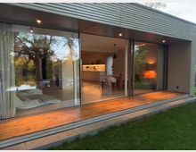 Modernes Wohnhaus mit großer Glasfront, die einen Blick ins helle Wohnzimmer und die offene Küche ermöglicht. Außen eine Holzterrasse, umgeben von einem gepflegten Rasen und Bäumen im Hintergrund.