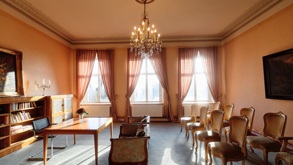 Eleganter Salon- oder Besprechungsraum mit hohen Decken, Stuckrand und zentralem Kronleuchter. Drei große Fenster mit langen Vorhängen lassen viel Tageslicht herein; rechts stehen mehrere gepolsterte Stühle in Reihen, links ein Tisch mit Stuhl sowie ein Bücherregal und ein Schrank.