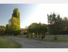 Parkanlage mit gepflastertem Weg, umgeben von Rasenflächen, Bäumen und Sträuchern. Im Hintergrund ist ein historisches Backsteingebäude mit roten Dachziegeln sowie ein Torbogen zu sehen.
