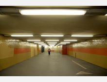 Ein unterirdischer Fußgängertunnel mit gelben und roten Fliesenwänden, beleuchtet durch Reihen von Deckenlampen. Im Hintergrund ist eine einzelne Person zu sehen, sowie eine Tür auf der rechten Seite und Wegmarkierungen auf dem Boden.