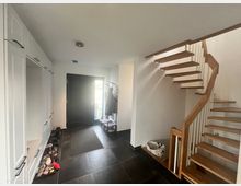 Eingangsbereich eines Hauses mit schwarzem Fliesenboden, einer schwarzen Haustür und weißen Wänden. Auf der rechten Seite führt eine Holztreppe mit offenen Stufen nach oben, links befinden sich weiße Einbauschränke, darunter stehen Schuhe.