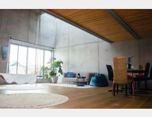 Helles Loft-Wohnzimmer mit hohen Betonwänden und schräger Holzdecke, großen Schiebetüren zur Terrasse und viel Tageslicht. Im Raum stehen eine freistehende Badewanne, Sofa mit Beistelltisch, Sitzsack, große Zimmerpflanze sowie ein Essbereich mit Tisch und Stühlen.