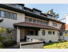 Wohnhaus im traditionellen Stil mit heller Putzfassade, rotem Ziegeldach und mehreren gitterförmigen Fenstern. Das Gebäude verfügt über einen überdachten Eingangsbereich und ist von Gartenpflanzen umgeben.