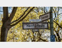 Straßenschilder einer Kreuzung mit den Namen „Herrfurthstr.“ und „Oderstraße“, montiert an einem grünen Metallpfosten. Im Hintergrund sind blühende Bäume und helle Frühlingsfarben erkennbar.