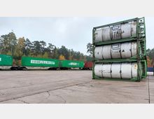 Ein Güterbahnhof mit gestapelten silbernen Tankcontainern in grünen Halterungen im Vordergrund und grünen Frachtcontainern auf einem Zug im Hintergrund. Der Bahnhof ist von einer Waldlandschaft mit herbstlich gefärbten Bäumen umgeben.