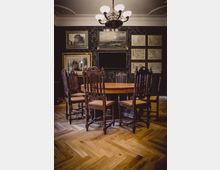 Ein elegantes Zimmer mit einem runden Holztisch und sechs kunstvoll geschnitzten Stühlen aus dunklem Holz. Die Wände sind mit schwarz gemustertem Tapeten bedeckt und dekoriert mit großformatigen Landschaftsgemälden, Karten und Bilderrahmen; an der Decke hängt ein Kronleuchter mit weißen Lampenschirmen.