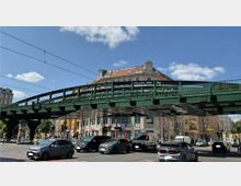 Eine grüne Stahlbrücke mit daran befestigten Oberleitungen überspannt eine Kreuzung mit fahrenden Autos. Im Hintergrund ist ein historisches Gebäude mit rotem Ziegeldach und großen Fenstern zu sehen, umgeben von weiteren Gebäuden und Geschäften.