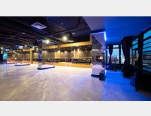 Moderner Innenraum einer Lounge oder eines Clubs mit glänzenden Spiegelwänden, Säulen mit Lichteffekten und einer erhöhten Sitzbereichsebene im Hintergrund. Große Fenster mit dunklen Vorhängen lassen den Blick nach draußen in eine Stadtlandschaft zu.