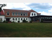 Zweistöckiges Gebäude mit weißer Fassade und roten Ziegeldächern, das auf der linken Seite mehrere Gaubenfenster besitzt. Davor befindet sich eine große Rasenfläche, und rechts ist ein überdachter Bereich mit Glasfronten zu sehen.