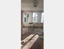 Ein verlassener Raum mit drei großen, schmutzigen Fenstern an einer weißen Wand, einer defekten Holzfußbodendiele, Schutt und verstreutem Müll am Boden. In der Mitte steht ein kleiner Tisch auf metallenen Beinen, darüber hängt eine rechteckige Deckenleuchte.