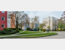 Wohnsiedlung mit mehrstöckigen Gebäuden in bunten Farben (Rot, Orange, Blau und Beige), umgeben von Gehwegen, Rasenflächen und Bäumen. Ein Radfahrer fährt auf einem Weg, und im Hintergrund sind Müllcontainer und parkende Autos sichtbar.