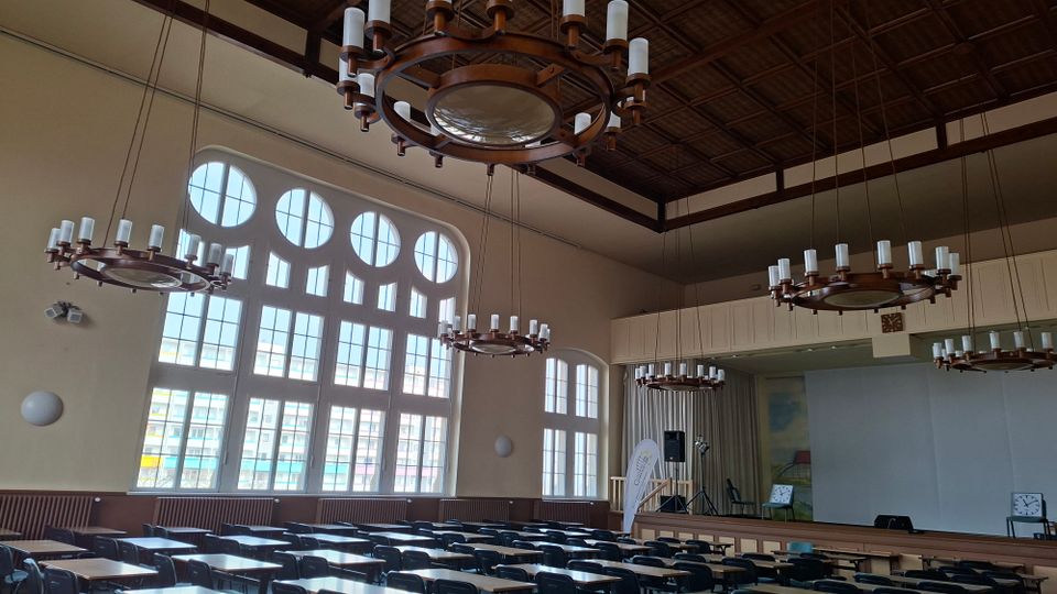 Großer Saal mit hohen, rundbogigen Sprossenfenstern und Holzbalkendecke, von mehreren ringförmigen Kronleuchtern beleuchtet. Im Vordergrund stehen viele Reihen von Tischen und Stühlen, hinten befindet sich eine Bühne mit Vorhang und Lautsprechertechnik.