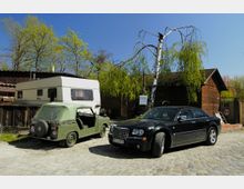 Ein kleiner Hof mit Kopfsteinpflaster, auf dem ein grünes Cabrio, ein schwarzer PKW und ein weißer Wohnwagen parken. Im Hintergrund stehen Bäume und ein Holzgebäude mit brauner Fassade.