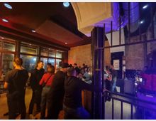 Innenraum einer Bar oder eines Cafés mit Backsteinwänden und großer Glasfront. Mehrere Personen stehen an einer Theke oder unterhalten sich in einer lockeren Atmosphäre bei gedimmtem Licht.