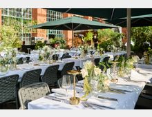 Terrasse eines Restaurants mit festlich gedeckten Tischen, dekoriert mit weißen Blumenarrangements und goldenen Kerzenhaltern, umgeben von grünen Pflanzen. Im Hintergrund ist eine Backsteinfassade mit großen Fenstern zu sehen.