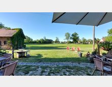 Garten- oder Hofbereich mit Blick auf eine weitläufige, gepflegte Wiese, umgeben von Bäumen. Links ein Backsteingebäude mit bewachsener Fassade, rechts ein Sonnenschirm und Terrassenmöbel, im Hintergrund mehrere Sonnenliegen auf der Wiese.