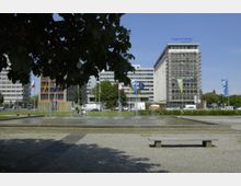 Öffentlicher Platz mit einem rechteckigen Brunnen und Wasserspielen im Vordergrund, umgeben von großflächigen Pflastersteinen, einer Sitzbank und Bäumen. Im Hintergrund stehen moderne Bürogebäude, darunter eines mit dem Schriftzug „HypoVereinsbank“, sowie mehrere wehende National- und Landesflaggen.