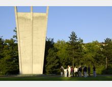 Ein großes, modernes Denkmal aus Beton mit zwei emporragenden Spitzen steht auf einer Wiese, umgeben von Bäumen; im Vordergrund ist eine Gruppe von Menschen, die das Denkmal betrachten. Der Himmel ist klar und blau.