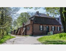 Backsteingebäude mit Ziegeldach und grünen Fensterläden, gelegen an einer gepflasterten Straße in einer parkähnlichen Umgebung mit Bäumen und Grünflächen. Im Hintergrund sind moderne Gebäude sichtbar.