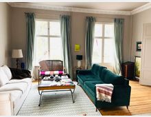 Wohnzimmer mit Holzboden, einem großen weißen Sofa und einem grünen Samtsofa. Vor den Sofas befindet sich ein niedriger Couchtisch, dahinter große Fenster mit hellgrünen Vorhängen, die viel Tageslicht einlassen.