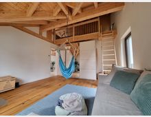 Wohnzimmer mit sichtbarer Holzbalkendecke und Zwischengeschoss, das über eine Holzleiter erreichbar ist. Der Raum ist hell, mit einem grauen Sofa, einer Hängematte in Türkis und einem Holzboden ausgestattet. Ein großes Fenster lässt Tageslicht herein.