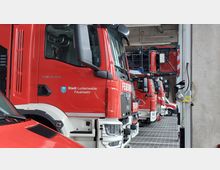 Mehrere rote Feuerwehrfahrzeuge der "Stadt Luckenwalde Feuerwehr" stehen geparkt in einer modernen Fahrzeughalle mit Betonwänden und grauem Fliesenboden. Im Vordergrund ist ein großer Löschwagen sichtbar, dahinter weitere Einsatzfahrzeuge in einer Reihe.
