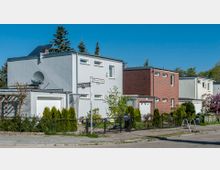 Reihenhäuser in einem Wohngebiet, bestehend aus modernen Gebäuden mit flachen Dächern und hellen Fassaden, darunter Putz und Ziegel. Vorgärten mit Rasen, Sträuchern und Zäunen prägen die Umgebung, daneben Straßenschilder und ein Gehweg.