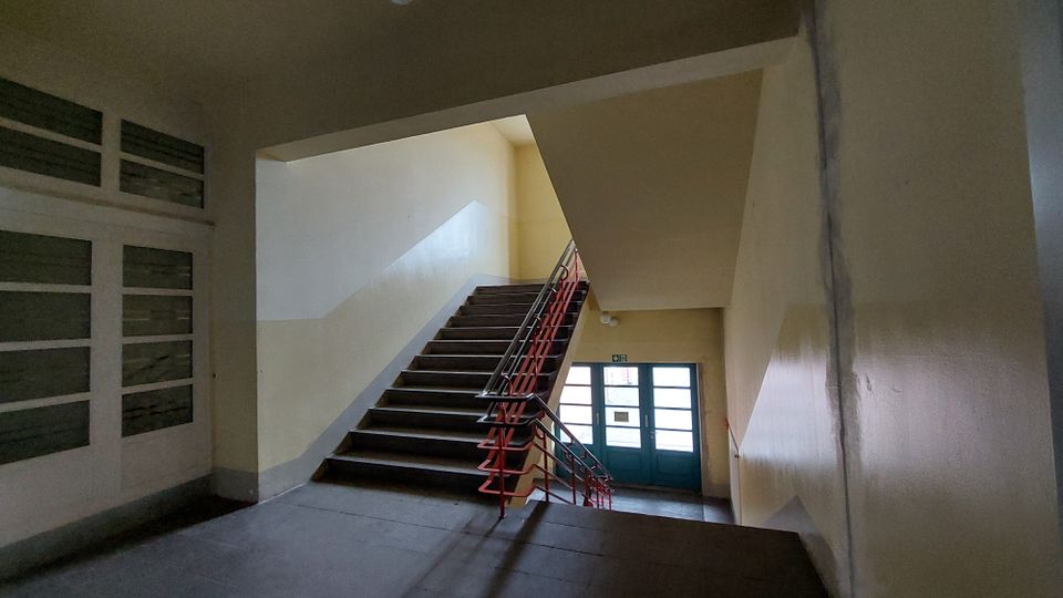 Treppenhaus eines Gebäudes mit beige gestrichenen Wänden, dunkelbraunen Stufen und einem roten Geländer. Im Hintergrund befindet sich eine Tür mit mehreren Glasfeldern, die zu einem Ausgang führt.