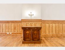 Holzvertäfelter Innenraum mit breiten Dielenboden und weißen Wänden. In der Mitte steht ein dunkles Holzpult mit gedrechselten Säulen, darüber eine Wandlampe im klassischen Stil.