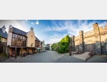 Alt-Text: Eine gepflasterte Gasse in einem historischen Themenbereich mit Fachwerk- und Steingebäuden. Links stehen Häuser mit Holzfenstern und Dachschindeln, rechts eine massive Steinmauer mit Holztoren und einer Zugbrücke, von Pflanzen bewachsen.