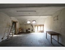 Großer, leerer Dachbodenraum mit schrägen Decken, abgenutztem Holzboden und fleckigen, teils rissigen Wänden. Hinten sorgen kleine Dachfenster und ein größeres Fenster für Licht; im Raum stehen eine Leiter, ein Tisch sowie einige alte Möbel.